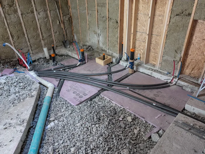 Underfloor rough-in with PEX manifold lines and drain stubs set before a basement slab is poured